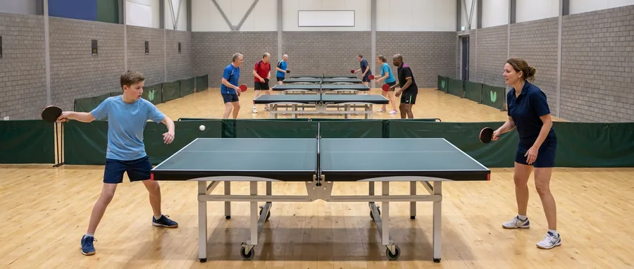 Een jonge speler slaat een backhand aan de tafel terwijl de trainster aan de overkant toekijkt